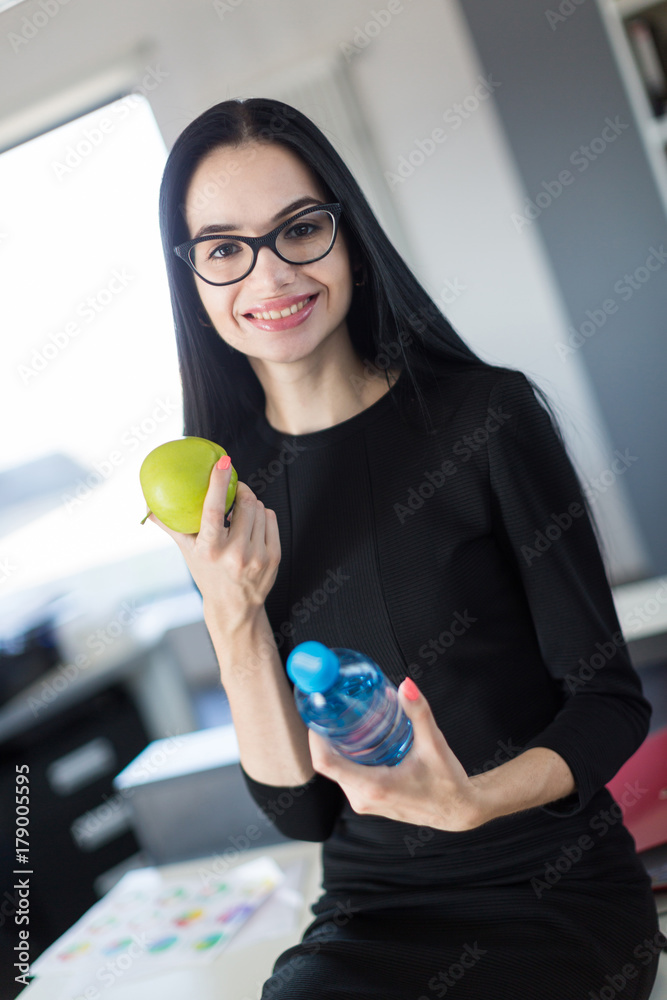 Beautiful young businesswoman in black dress and glasses sit on table in the office and hold green apple and bottle Beautiful young businesswoman in black dress and glasses sit on table in the office and hold green apple and bottle