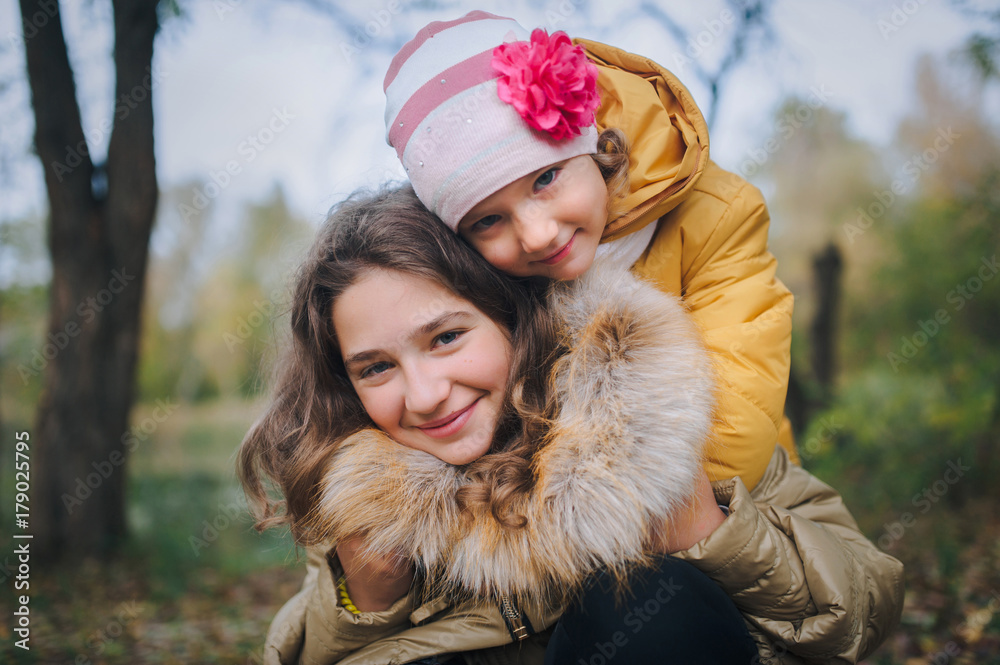 Happy smiling children are having fun in the autumn park. The younger sister hugs her older sister. Happy smiling children are having fun in the autumn park. The younger sister hugs her older sister.
