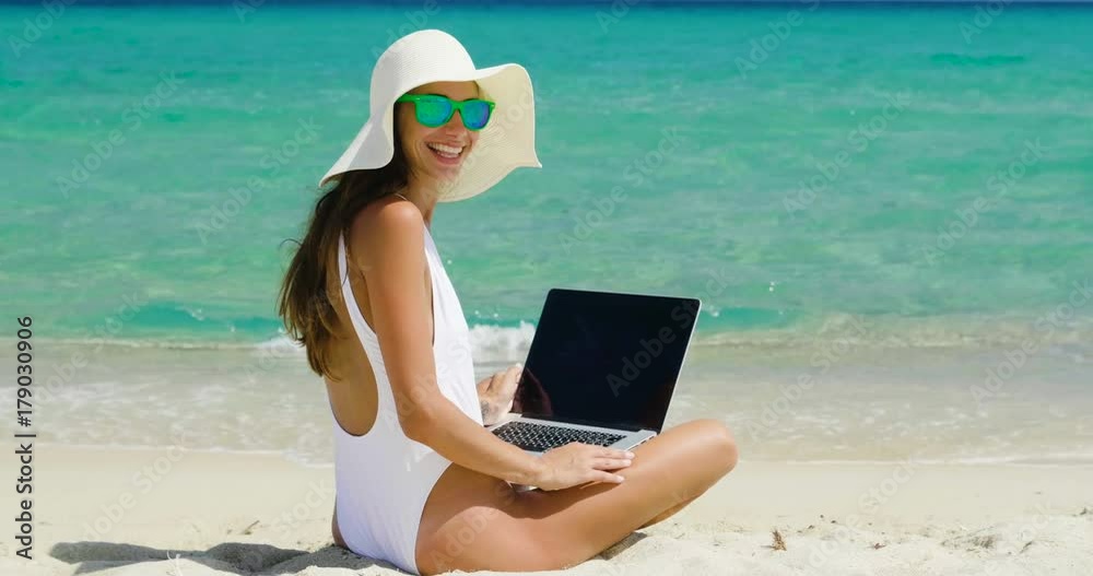 A beautiful woman on holiday with the white costume and the straw hat on the shore of a beautiful sea uses the computer to work or shop. Concept of: vacation, work, shopping,... A beautiful woman on holiday with the white costume and the straw hat on the shore of a beautiful sea uses the computer to work or shop. Concept of: vacation, work, shopping,...