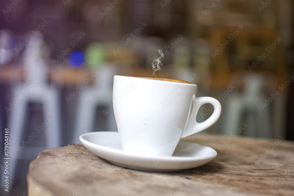 White cup with steaming hot cappuccino on the wooden  table with blurred coffee bar background
