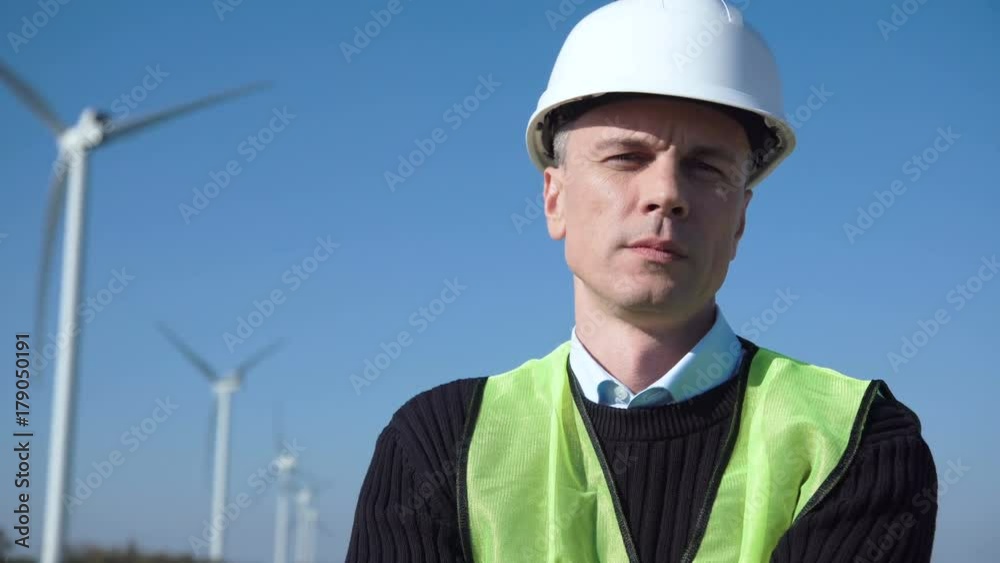 Close shot of adult engineer in green vest and white hard hat with wind turbines on background looking at camera Close shot of adult engineer in green vest and white hard hat with wind turbines on background looking at camera