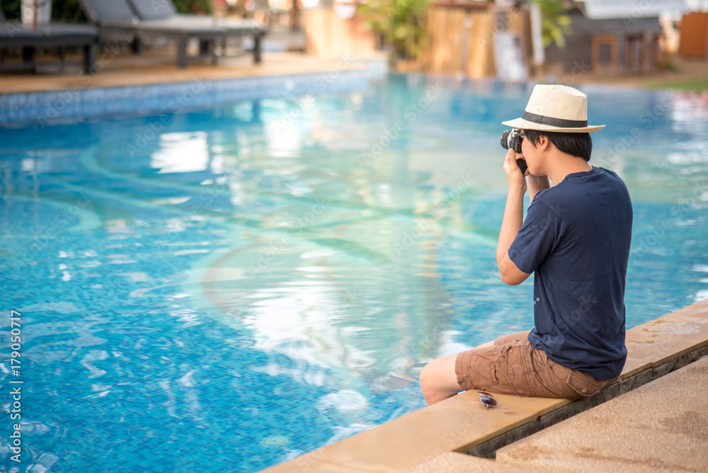 Young Asian man sitting on the edge of swimming pool and taking photo with his camera at resort, relaxing time and summer holiday vacation concepts