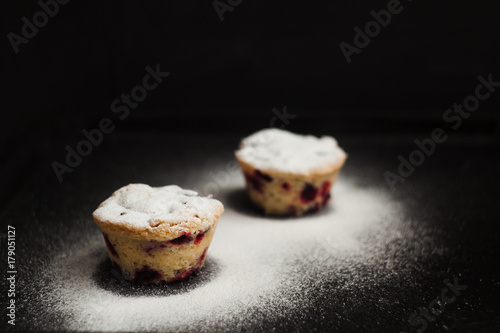 Close up of a delicious berry muffins on a dark table, against black background, with snow like powder sugar scattered around and falling above