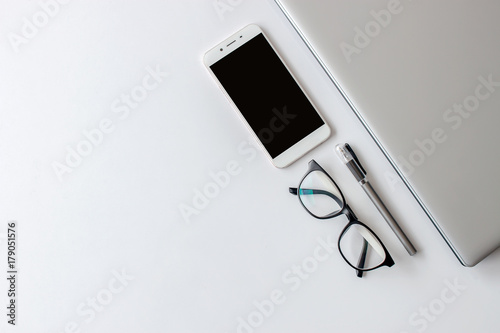 white desk table with office supplies notebook computer, smartphone and eyeglasses, top view, over light and  soft-focus in the background photo