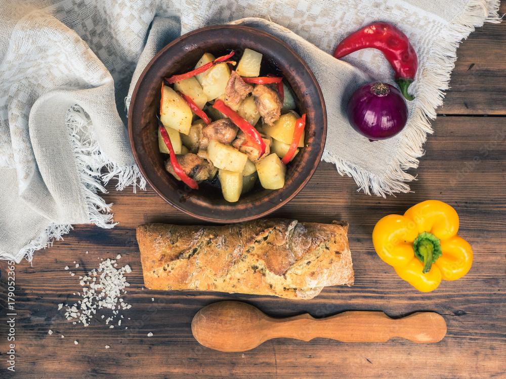 Rural still life. Baked meat with potatoes, bread, salt, pepper and onion on the background of wooden table and coarse cloth. Roast potatoes in a clay bowl with a wooden spoon. Top view. Rural still life. Baked meat with potatoes, bread, salt, pepper and onion on the background of wooden table and coarse cloth. Roast potatoes in a clay bowl with a wooden spoon. Top view.