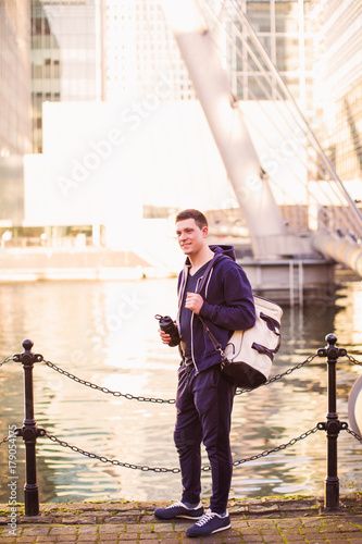 The young sporty man standing outdoor by the river and holding sporty bag and bottle