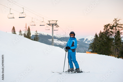Young beautiful female on ski holiday in mountains looking at the camera and smiling. Woman at ski resort wearing helmet, blue ski suit and goggles.