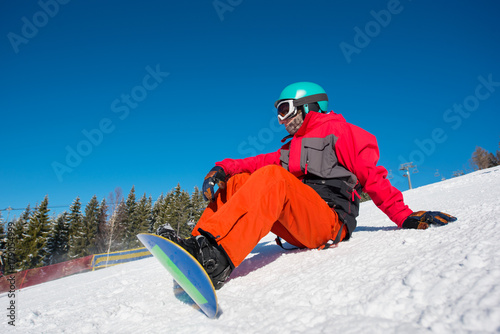 Male snowboarder sitting, relaxing on the slope of the hill at winter ski resort. Blue sky, forest on the background. copyspace lifestyle activity hobby sportsman recreation travelling Bukovel
