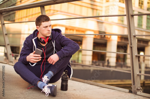 The young sporty man sitting on the floor outdoor and holding the phone in hand and looking aaside