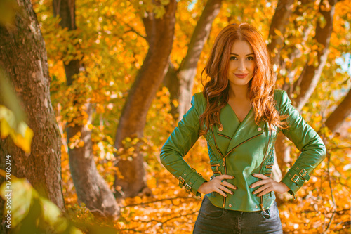 Beautiful and nice red-haired lady, looks stylish, dressed in a green leather jacket and black jeans. A successful combination of colors and women's casual wardrobe, a daily winning look