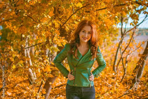 Beautiful and nice red-haired lady, looks stylish, dressed in a green leather jacket and black jeans. A successful combination of colors and women's casual wardrobe, a daily winning look