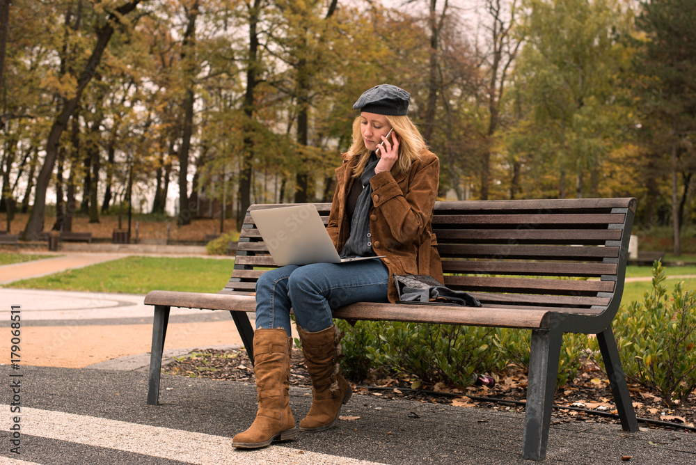 Beautiful young woman is sitting on the bench in the park on an autumn day and she is working on her laptop Beautiful young woman is sitting on the bench in the park on an autumn day and she is working on her laptop
