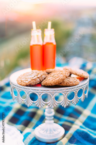 The close-up vertical photo of the white board with cookies and two glass bottles with orange juice in the mountains during the sunset. photo