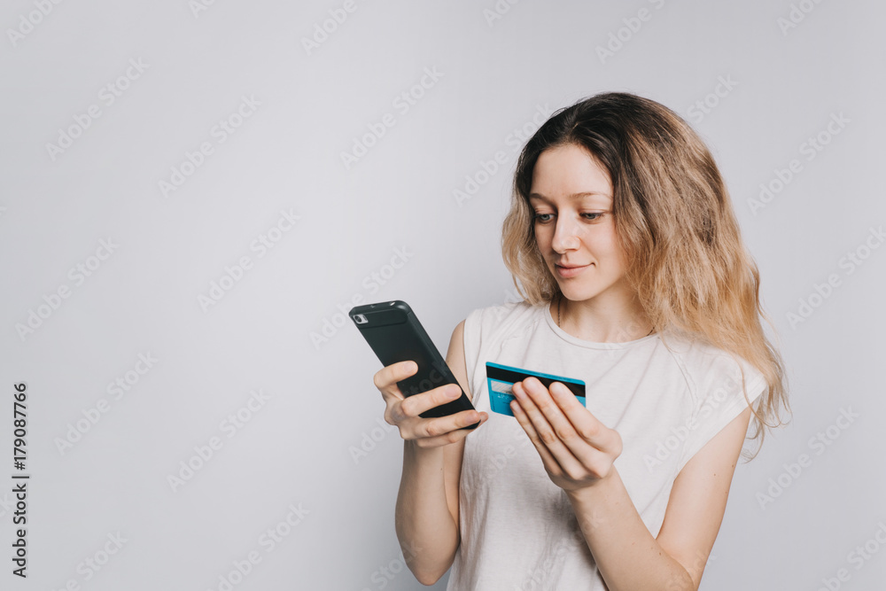 young beautiful Caucasian girl in a white t-shirt makes a purchase using a mobile phone and credit card on white background. Enters a credit card number young beautiful Caucasian girl in a white t-shirt makes a purchase using a mobile phone and credit card on white background. Enters a credit card number