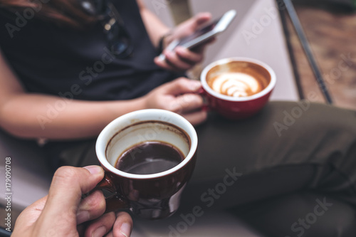 Closeup image of a man's hand holding black coffee cup with a woman using smartphone while drinking coffee in cafe