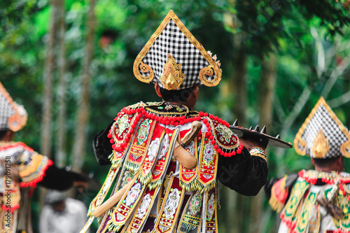 Men in traditional costumes perform a Balinese war dance called Tari Baris Gede outdoors. Back view.