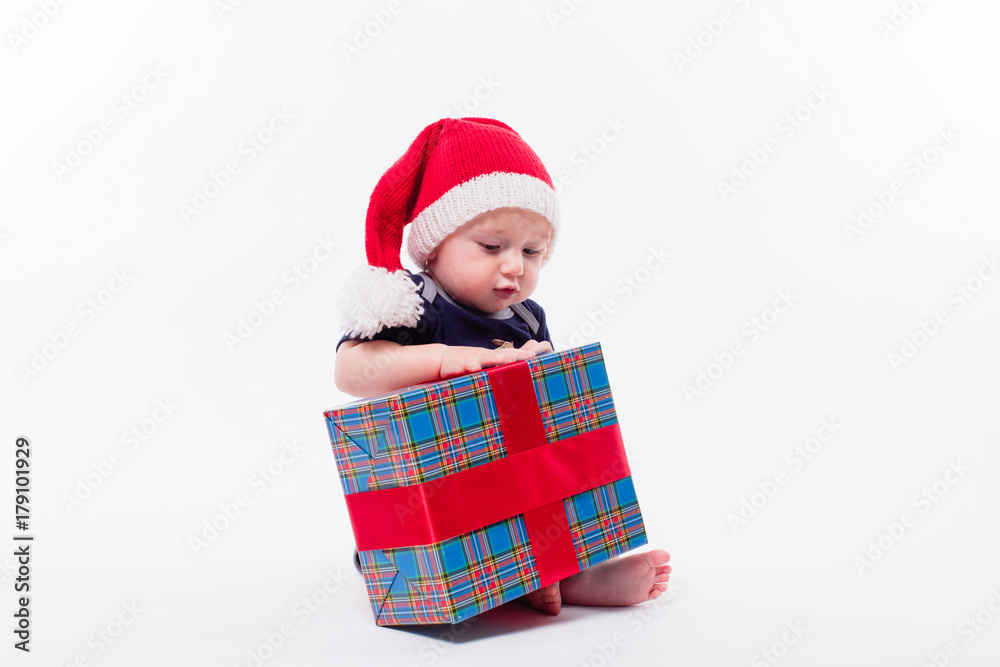 cute toddler is sitting in a New Year's red cap on white background unpacks a gift with a smile on his face cute toddler is sitting in a New Year's red cap on white background unpacks a gift with a smile on his face