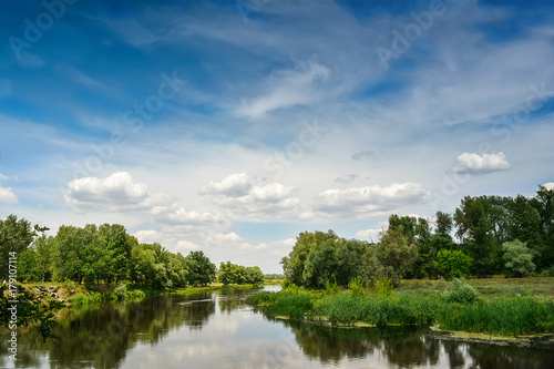 Beautiful sky with clouds and a river, surrounded by green trees. © Mykola