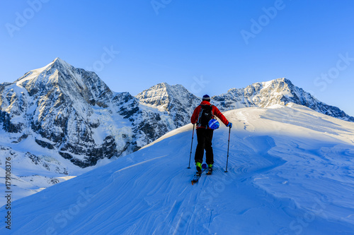 Mountaineer backcountry ski walking up along a snowy ridge with skis in the backpack. In background blue sky and shiny sun and Zebru, Ortler in South Tirol, Italy. Adventure winter extreme sport.