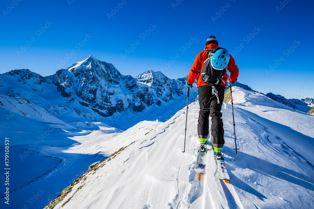 Mountaineer backcountry ski walking up along a snowy ridge with skis in the backpack. In background blue sky and shiny sun and Zebru, Ortler in South Tirol, Italy.  Adventure winter extreme sport.