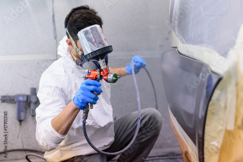 A varnisher is painting a bodywork of a car with a spray gun. No face but dust mask