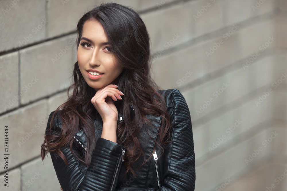 Fototapeta premium Portrait of beautiful smiling brunette, fashion and stylish standing near the wall. A curly hairstyle woman in total black look with leather jacket and dress.