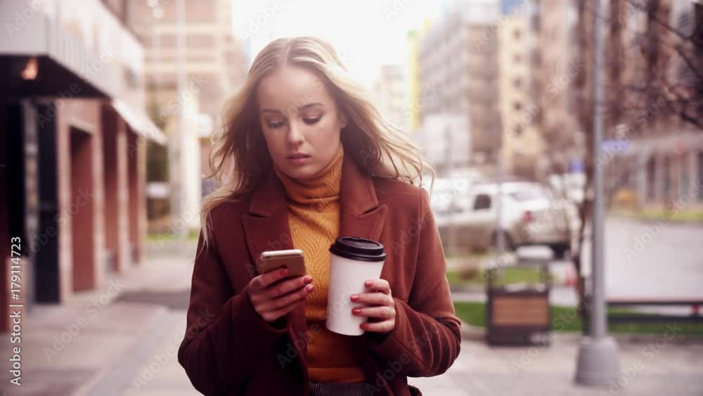 Stylish woman walking on street. Drink coffee and using smartphone.