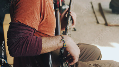 man playing the bass guitar at the concert in the street. Close up view of guitarist plays on brown wooden musical instrument. Street musician with a wicker hippie bracelet on his arm.