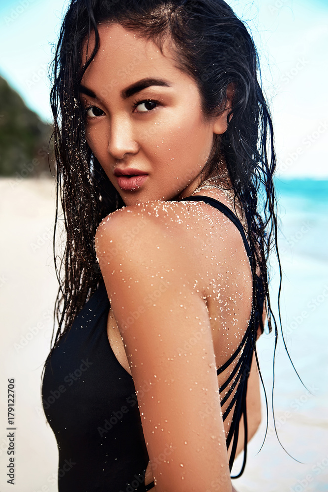 Fototapeta premium portrait of beautiful caucasian sunbathed woman model with dark long hair in dark swimsuit posing on summer beach with white sand on blue sky and ocean background