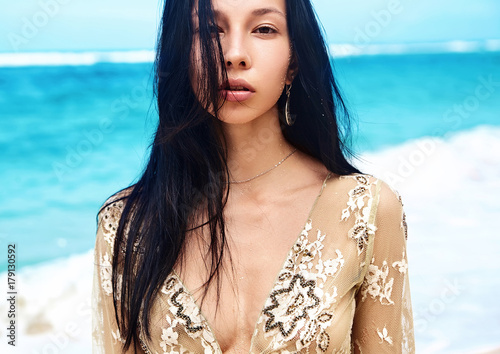 sensual portrait of beautiful caucasian woman model with dark long hair in beige blouse posing on summer beach with white sand on blue sky and ocean background photo