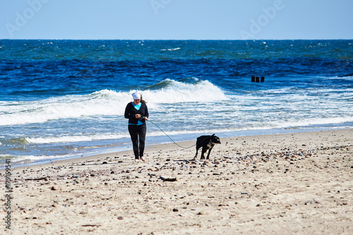 Kobieta spaceruje po plaży w wietrzny dzień z dużym czarnym psem.