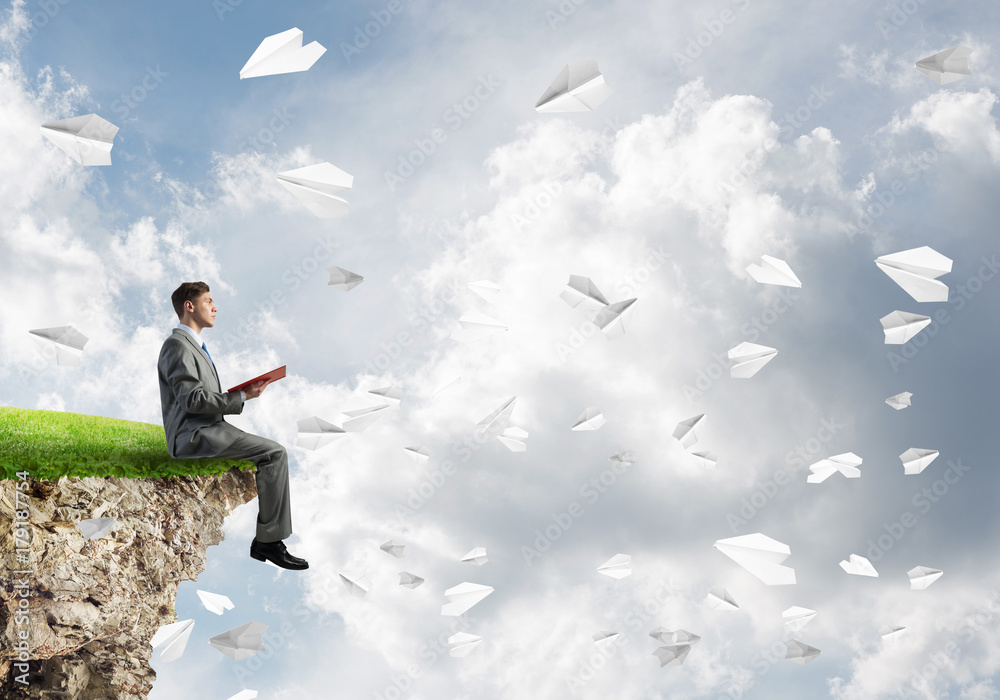Man on edge reading book and paper planes flying in air Man on edge reading book and paper planes flying in air