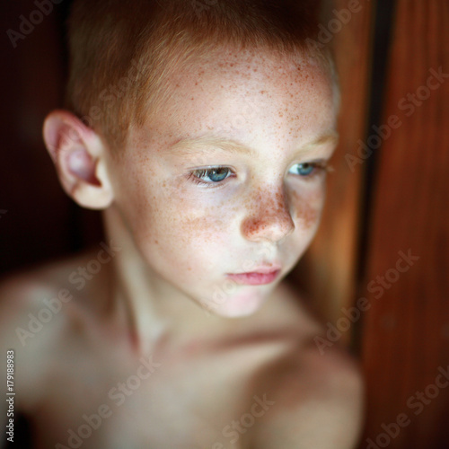 Portrait of young boy no shirt, red hair, blue eyes & freckles. His head is turned to the side showing a three quarter view of face.