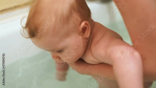 Cute baby boy taking bath mother hands pouring water back child half year old infant kid health care chubby cheeks beautiful big blue eyes looking camera closeup enjoying bathing wash clean skin body photo