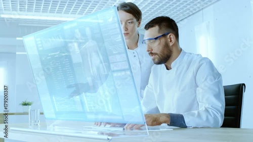 In the Futuristic Laboratory Male and Female Scientists Work on Transparent Computer Display, They Try to Prolong Human Life. Screen Shows Various Human Related Infographics and Data. 4K UHD.