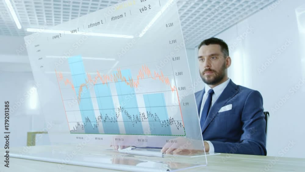 In the Near Future Successful Businessman Works on Computer with Transparent Display that Shows Interactive Line Charts and Other Useful Information. His Office is Bright and Modern. 