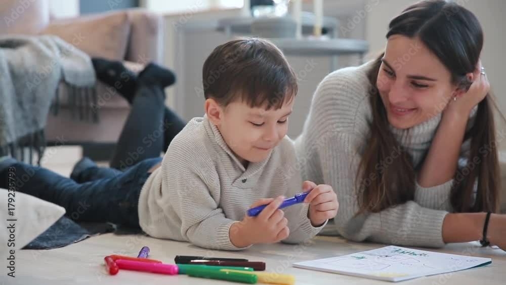 Beautiful young mother in a warm sweater lying on the floor with my son drawing with markers on paper portraying his family. The child learns to draw. Close-up Beautiful young mother in a warm sweater lying on the floor with my son drawing with markers on paper portraying his family. The child learns to draw. Close-up