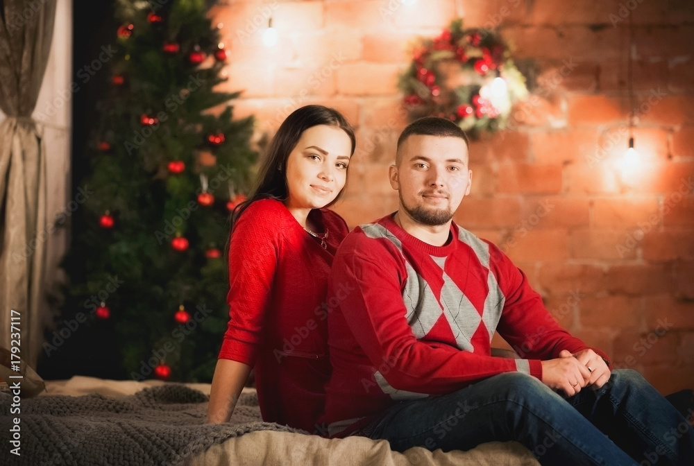 A man and a woman are hugging on the bed against the background of a Christmas tree. Beautiful happy young family celebrating Christmas together. Lovers on the New Year together in an apartment A man and a woman are hugging on the bed against the background of a Christmas tree. Beautiful happy young family celebrating Christmas together. Lovers on the New Year together in an apartment