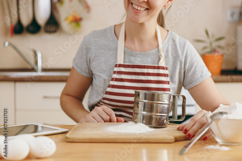 A young smiling woman with iron sieve and flour on the table in the kitchen. Cooking home. Prepare food.