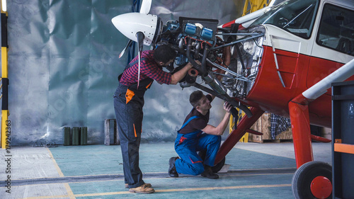 Two mechanics working on a small aircraft in a hangar with the cowling off the engine as they perform a service or repair photo