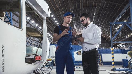 Aircraft mechanic and draftsman standing near plane carcass and talking with each other.
