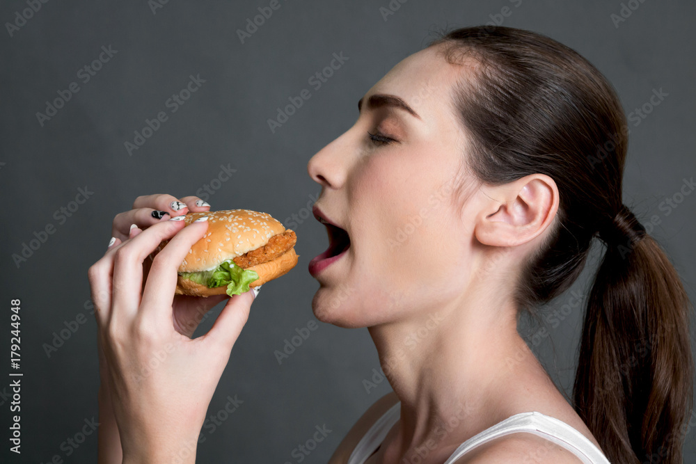 Young woman eating hamburger on gray background. Junk food and fast food concept Young woman eating hamburger on gray background. Junk food and fast food concept