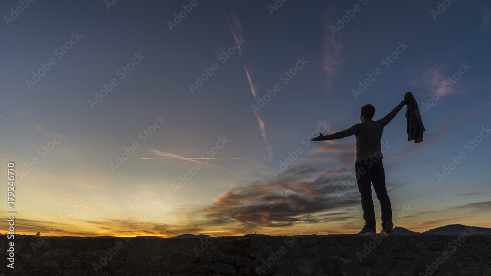 Man in casual clothes holding his sweater standing on a wall against a sunrise