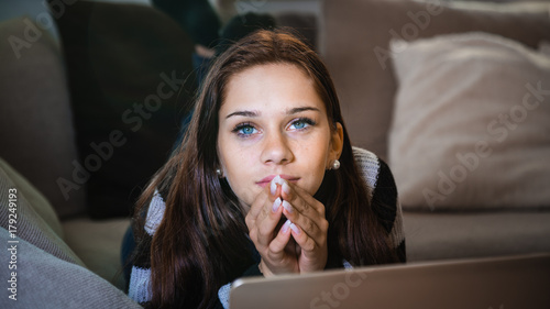 Portrait of an attractive young brunette woman lying down on her stomach on sofa looking at the laptop monitor with hands on chin