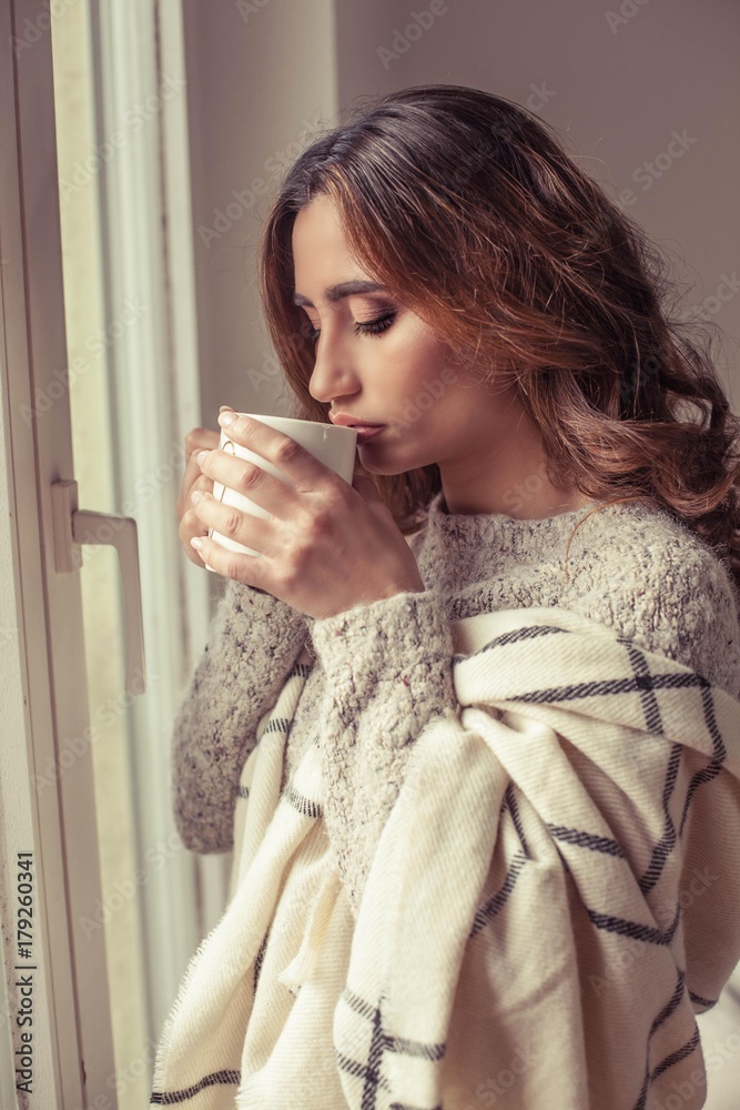 Young beautiful woman stands near a window, wrapped in a blanket and drink hot coffee. Cozy. Autumn. Warm cozy evening. Young beautiful woman stands near a window, wrapped in a blanket and drink hot coffee. Cozy. Autumn. Warm cozy evening.