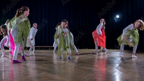 Girls are wearing white costumes and capes of different colours and are performing in a school play and are dancing hip hop and modern dancing.