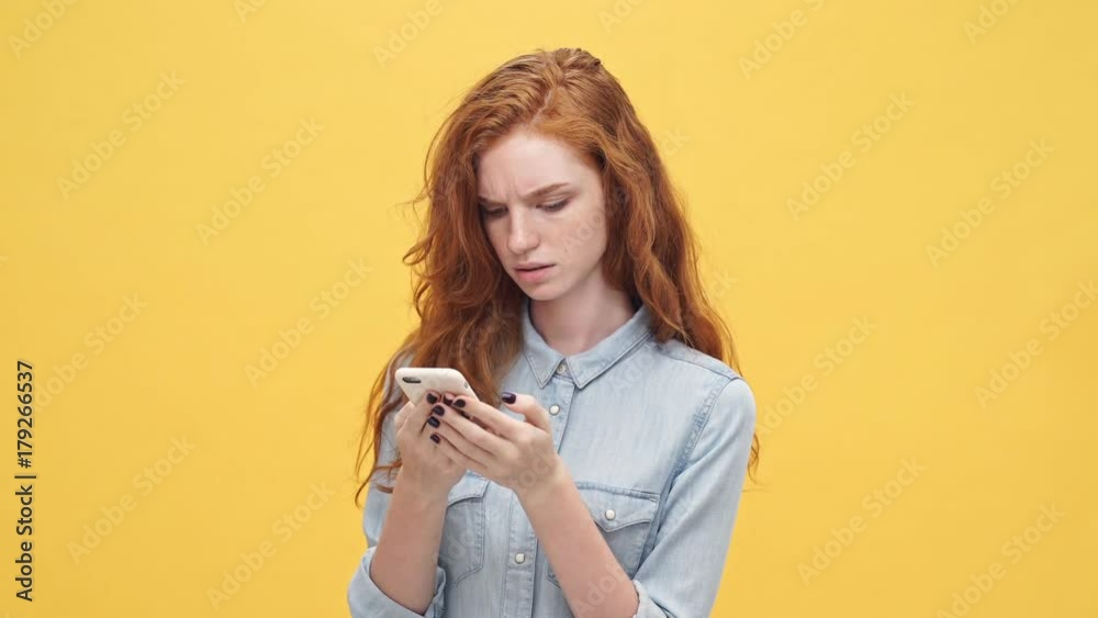 Displeased ginger woman in denim shirt writing message on smartphone over yellow background Displeased ginger woman in denim shirt writing message on smartphone over yellow background