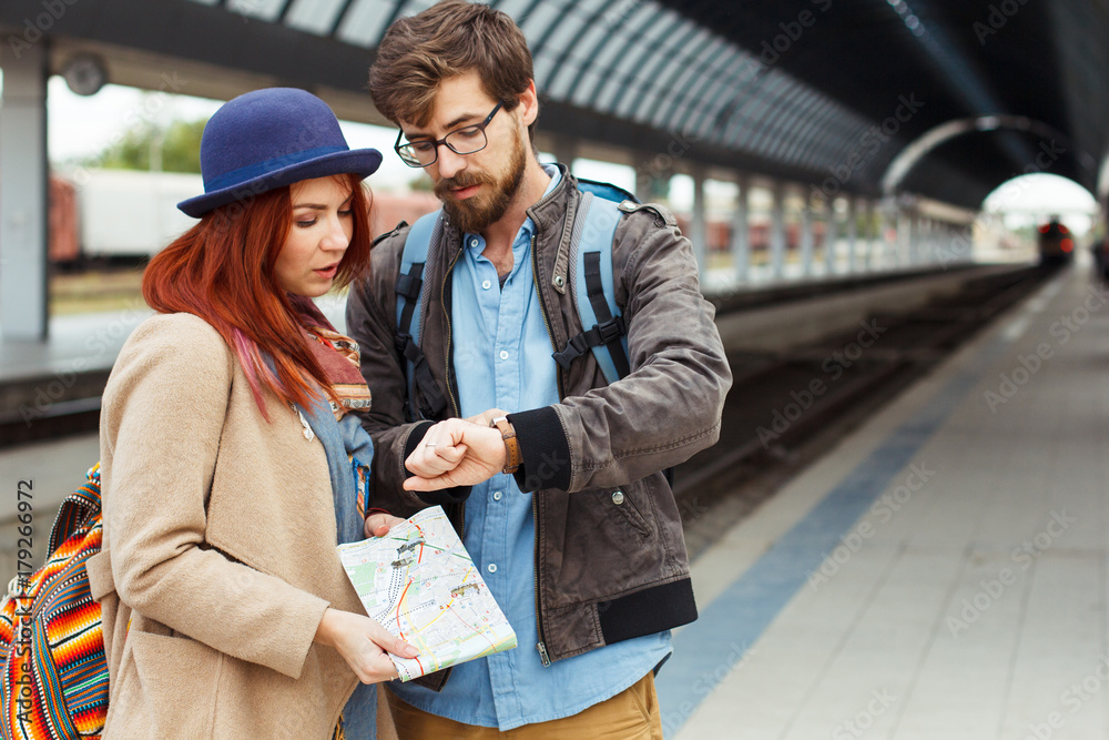 Hipster Traveller couple looking at smart watch while waiting for the train at railway station. Autumn time. Woman holding a map. Man with backpack. Copy space. Travel concept