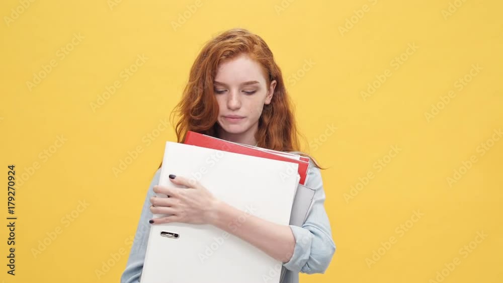 Young ginger female worker in denim shirt holding many folders and listening instructions over yellow background Young ginger female worker in denim shirt holding many folders and listening instructions over yellow background