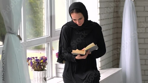 Beautiful meddle eastern young woman reading book next to window.